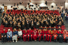 Duża grupa osób w czarnych togach akademickich stoi w audytorium i wyrzuca w górę birety z pomarańczowymi chwostami, świętując zakończenie uroczystości. W pierwszym rzędzie siedzi kilkanaście osób w czerwonych togach z ozdobnymi łańcuchami, prawdopodobnie przedstawiciele władz uczelni, obok nich kilka osób w strojach cywilnych.