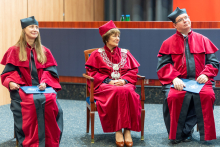 Trzy osoby: władze dziekańskie i prorektorka, ubrani w czerwone togi i czerwone birety. Siedzą na środku auli i słuchają przemówienia.