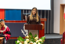 Młoda, elegancko ubrana kobieta stoi przy mównicy i przemawia. Przed mównicą widać bukiet kwiatów. W tle ekran multimedialny i kolorowe banery.