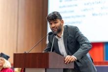 Młody, elegancko ubrany mężczyzna przemawia za mównicą. Podbiera się o mównicę dwiema rękami. Za nim widać ekran multimedialny i kolorowe banery.