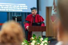 Mężczyzna w średnim wieku, w okularach, ubrany w togę akademicką w kolorach czerwonym i czarnym i w czarnym birecie stoi za mównicą i przemawia. Przed mównicą widać bukiet kwiatów. Za przemawiającym widać ekran z prezentacją multimedialną.