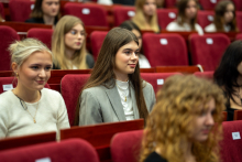 Kilka osób siedzi w czerwonych, miękkich fotelach w auli. Na pierwszym planie widać osobę w jasnoszarej marynarce i białej bluzce, obok siedzi osoba w białym swetrze. W tle znajdują się kolejne rzędy zajętych miejsc.