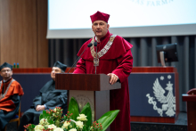 Prorektor w bordowej todze i birecie stoi przy drewnianej mównicy i przemawia. W tle siedzą dwie osoby w ciemnych togach, widać też fragment ekranu z logo wydziału.