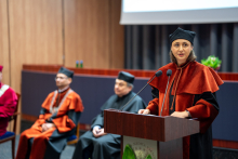 Prodziekan ds. nauczania na kierunku analityka medyczna, w czarno-pomarańczowej todze, stoi przy mównicy i przemawia. W tle siedzą trzy osoby w togach.