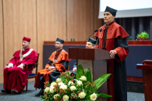 Prodziekan ds. nauczania na kierunku farmacja, w czarno-pomarańczowej todze, stoi przy mównicy i przemawia. W tle siedzą trzy osoby w togach.