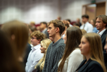 Studenci w czasie składania przysięgi. Są to osoby młode, nieformalnie ubrane. Wyglądają na skupione. Patrzą przed siebie. Stoją w równych rzędach w auli.
