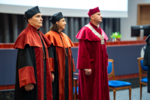 Trzy osoby, prorektor i prodziekanki, ubrani w togi akademickie, stoją na środku audytorium. Są skupieni i poważni. W tle widać krzesła, mównicę i ekran multimedialny.
