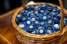 Wiklinowy okrągły kosz pełen srebrnych przypinek. Każda przypinka zapakowana jest w opakowanie z przezroczystym wieczkiem i granatowym spodem. Na przypinkach jest logo Wydziału Farmaceutycznego.