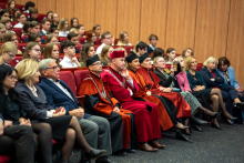 Kilka rzędów osób siedzi w auli na czerwonych fotelach. W pierwszym rzędzie siedzą osoby w bordowych i pomarańczowych togach oraz osoby w eleganckich strojach. Wszyscy patrzą przed siebie, twarze mają skupione. Słuchają wykładu.