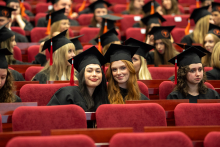 Absolwenci w togach i biretach siedzą w audytorium; na pierwszym planie dwie osoby uśmiechają się do kamery.