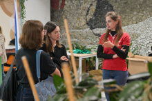 Three people are standing in a room, talking. They are dressed casually. In the background, there is a mosaic wall and several tables.