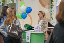 Four people at a green information stand. They are talking.  Green materials are lying on the counter, and next to it is a bouquet of green, blue, and white balloons. 