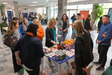 A dozen or so people in a bright hall with marble flooring. In the center stands a table with promotional materials, cans of drinks, and a basket of oranges. Several people are talking at the table, one is handing out flyers. Others are standing around. Plants and advertising banners are visible in the background.