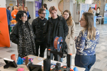 Five people standing at a table with a 3D printer and colorful figure-shaped prints.  In the background, there are stands with banners and people in orange sweatshirts.