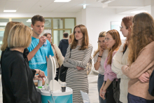 The photo shows seven people standing at a small stand in a bright room. They are talking. 