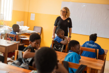 Kilka osób w sali lekcyjnej. Kobieta, dr hab. Joanna Peradzyńska, stoi przy drewnianych ławkach, przy których siedzą dzieci, wszystkie ubrane w takie same niebieskie koszulki. Większość dzieci patrzy na kobietę, jedno jest odwrócone przodem do zdjęcia.  