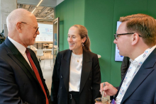 trzy osoby stoją w rozmowie w nowoczesnym wnętrzu z zieloną ścianą. Jedna osoba trzyma kubek z łyżeczką, a na szyi ma zawieszoną smycz z identyfikatorem. 