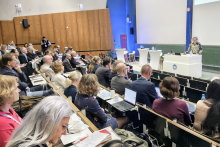 Na zdjęciu widać salę wykładową wypełnioną uczestnikami konferencji siedzącymi w rzędach. Na stołach znajdują się laptopy, notatniki, długopisy i kubki.