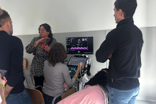 Five people in a bright room with an ultrasound machine. One person is lying on a treatment bed covered with green fabric, and another is sitting at the machine and operating it.