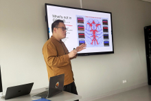 A man standing in a room in front of a large screen. The person presents a slide titled “What's not in GRASS...” with a colorful diagram of blood vessels and tables on both sides. There are two laptops on the table in front of the person.
