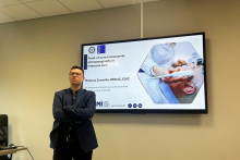 A man standing in a conference room next to a large screen. The screen displays a slide titled “Point-of-care transcranial ultrasonography in intensive care” and a photo of a person undergoing a head ultrasound. The speaker's name and institutional logos appear below the title.