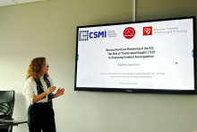 A woman standing in a conference room next to a large screen. The screen displays a presentation slide titled “Neurocritical Care Monitoring in the ICU: The Role of Transcranial Doppler (TCD) in Assessing Cerebral Autoregulation.” 