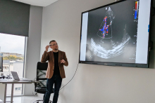 A man standing in a room in front of a large screen. The person points to an image displayed on the screen, which shows a color Doppler ultrasound image of the heart. 