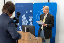A man standing at a lectern, holding a microphone and some papers. Two blue banners in the background. On the right, another man operating a camera on a tripod.