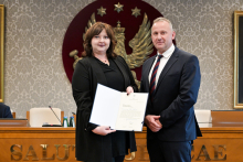 Dwie osoby, kobieta i mężczyzna, stoją obok siebie w formalnym otoczeniu. Osoba po lewej stronie trzyma otwarty dokument skierowany w stronę aparatu.