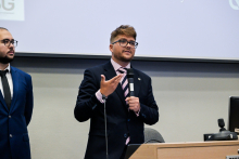 Dwaj mężczyźni w garniturach stoją przed publicznością. jeden z nich trzyma mikrofon i gestykuluje ręką, drugi stoi obok i patrzy w stronę uczestników.