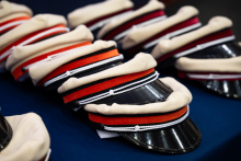 Na ciemnoniebieskim stole leżą rzędy kremowych czapek z czarnymi daszkami. Czapki mają opaski w kolorach: czerwonym, pomarańczowym, czarnym i bordowym. 