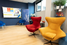 Jasny pokój. Na pierwszym planie stoją czerwony i żółty fotel. W tle widnieje ekran z napisem „Welcome Center at WUM” oraz plakat z logo uczelni. Po lewej stoją niebieskie krzesła, po prawej szafka z kolorowymi kubkami. Pokój ma drewnianą podłogę i duże okno.