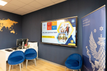 Jasny pokój. Po lewej widać białe biurko z niebieskimi krzesłami, po prawej rollup WUM. Na ścianie wisi ekran z informacjami o Welcome Center.
