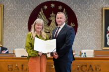 Dwie osoby, kobieta i mężczyzna, stoją obok siebie w formalnym otoczeniu. Osoba po lewej stronie trzyma otwarty dokument skierowany w stronę aparatu.