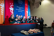 Na scenie stoi jedenaście osób – kobiety i mężczyźni – i śpiewa z otwartych nut. Przed nimi dyrygent w ciemnym garniturze prowadzi chór. W tle wiszą kolorowe banery z nazwą uczelni i hasłem inauguracji. Na stole przed sceną leżą czapki akademickie. Po prawej stronie siedzi publiczność.