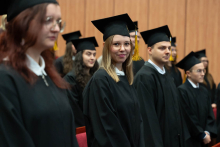 Grupa absolwentów ubrana w czarne togi stoi w rzędach w auli. Jedna z absolwentek patrzy wprost na aparat, uśmiecha się.