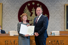 Dwie osoby, kobieta i mężczyzna, stoją obok siebie w formalnym otoczeniu. Osoba po lewej stronie trzyma otwarty dokument skierowany w stronę aparatu.