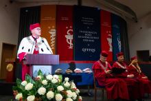 Rektor w czerwonym birecie i czerwonym płaszczu z białym kołnierzem przemawia przy mównicy. Za nim siedzą trzy osoby w czerwonych togach i biretach. W tle wiszą kolorowe banery z nazwami wydziałów i napisem „Inauguracja Roku Akademickiego 2025/2026”. Przed mównicą stoi bukiet białych róż z czerwonymi akcentami.