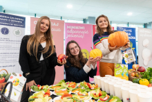 Trzy osoby stoją za stołem z talerzykami pełnymi świeżych warzyw i kubkami. Kobieta po lewej stronie trzyma jabłko, osoba w środku trzyma małą dynię, a kobieta po prawej stronie trzyma dużą dynię