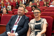 Kobieta i mężczyzna siedzą obok siebie na czerwonych fotelach podczas konferencji. Mężczyzna ma garnitur i identyfikator, kobieta jasną sukienkę z czarnymi wzorami i okulary. W tle inni uczestnicy.