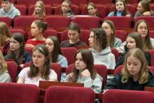 A group of young people in red chairs in the auditorium listen attentively to the lecture.