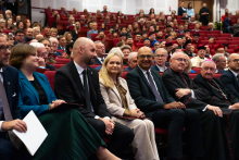 W audytorium siedzi duża grupa osób. W pierwszym rzędzie znajduje się sześć osób, w tym kobieta w jasnym garniturze i mężczyzna w ciemnym garniturze. W tle widać pełną salę z czerwonymi fotelami. Większość osób nosi eleganckie ubrania, część ma togi akademickie.