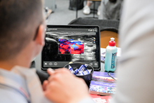 Two people are looking at a tablet screen displaying a color ultrasound image. On the table next to them is a bottle of ultrasound gel and a bowl with small items. Blurred silhouettes of people can be seen in the background.