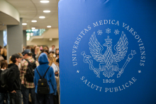 Na pierwszym planie stoi duży niebieski baner z białym logo WUM i napisem „Universitas Medica Varsoviensis Saluti Publicae 1809”. W tle widać kilkanaście osób w korytarzu, część z nich rozmawia przy stoiskach