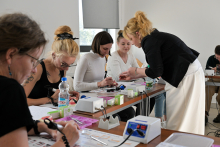 Grupa kobiet siedzi przy stole w jasnym pomieszczeniu, uczestnicząc w warsztatach. Na stole znajdują się butelki z wodą, pudełka z przyborami oraz urządzenia elektroniczne