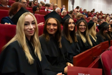 Młode kobiety, ubrane w czarne togi i birety. Siedzą na czerwonych fotelach w auli. Uśmiechają się do zdjęcia.