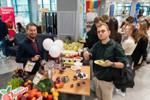 Grupa osób zgromadzona wokół stołu z owocami, warzywami i innymi produktami spożywczymi. 