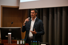 Mężczyzna w ciemnej marynarce i białej koszuli przemawia na scenie, trzymając mikrofon w ręce.