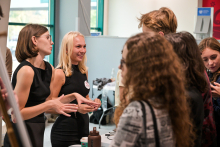 Kilka osób stoi przy stole w jasnym pomieszczeniu. Dwie osoby w czarnych sukienkach gestykulują i tłumaczą coś grupie. Na stole leżą małe przedmioty i butelka w brązowym kolorze. 