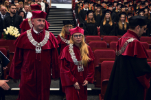 Osoby w bordowych togach akademickich z ozdobnymi srebrnymi łańcuchami ceremonialnymi idą między rzędami czerwonych foteli w audytorium. W tle widać dużą grupę osób w czarnych togach i biretach z pomarańczowym chwostem, stojących w rzędach na podwyższeniu. 